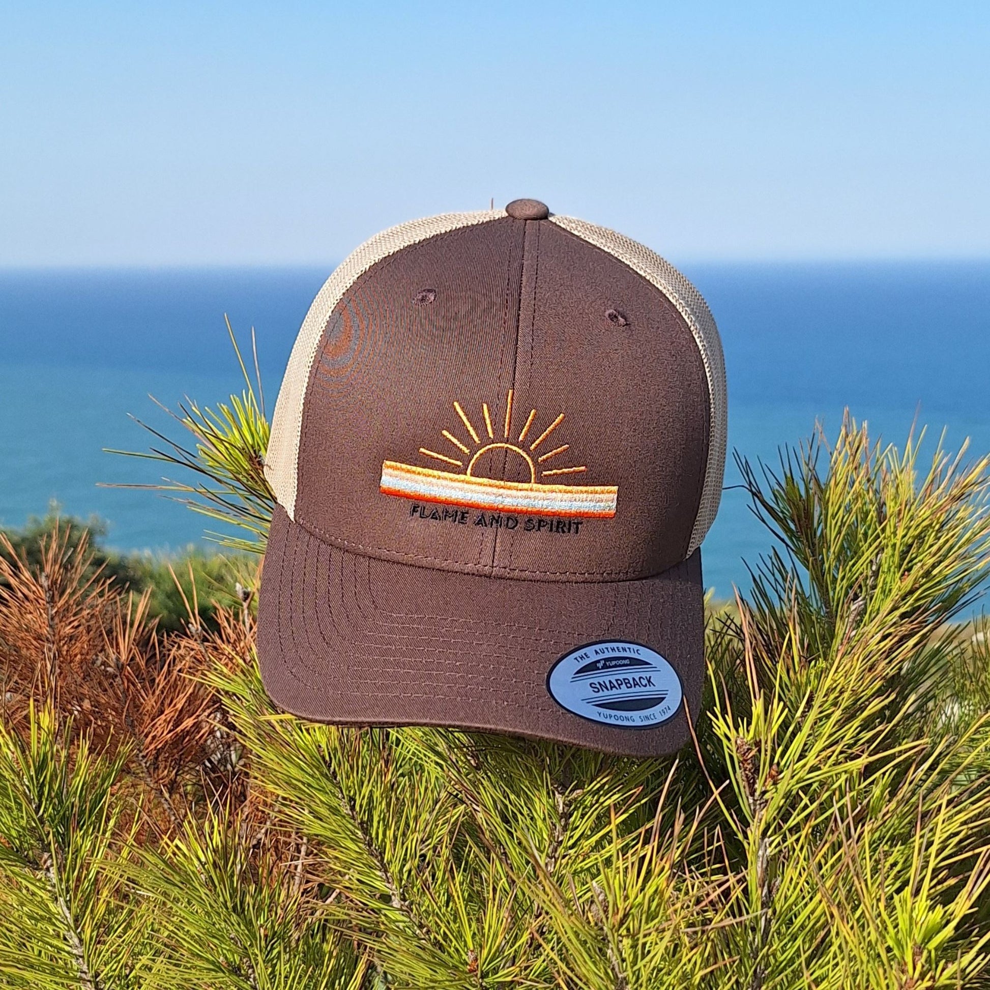 casquette trucker  non genrée, beige et marron portée par une femme avec un motif brodé soleil et bandes de couleurs ainsi que la marque flame and spirit brodée en noir