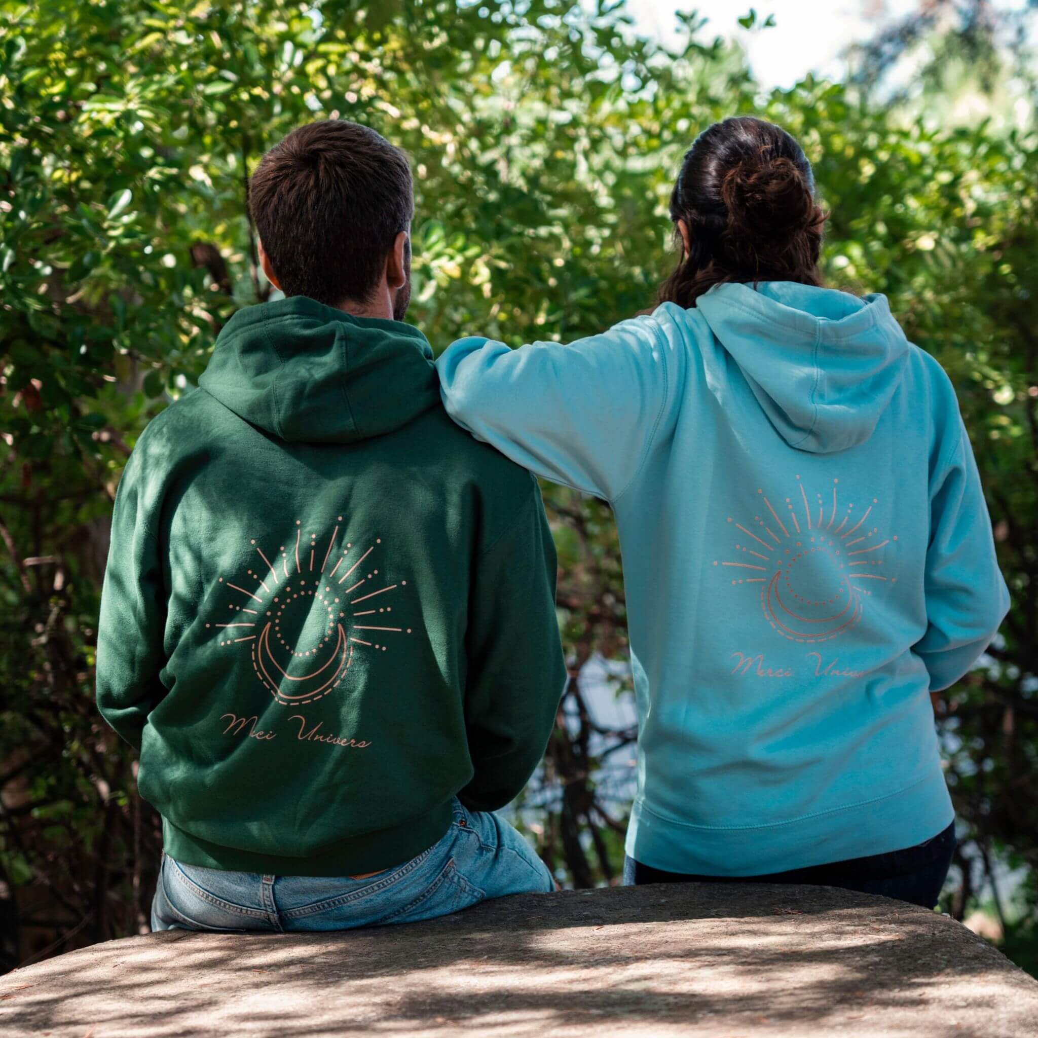 Deux personnes portent des sweats à capuche Flame & Spirit avec motif soleil et lune et message "Mon Univers", en coloris vert sapin et bleu ciel.