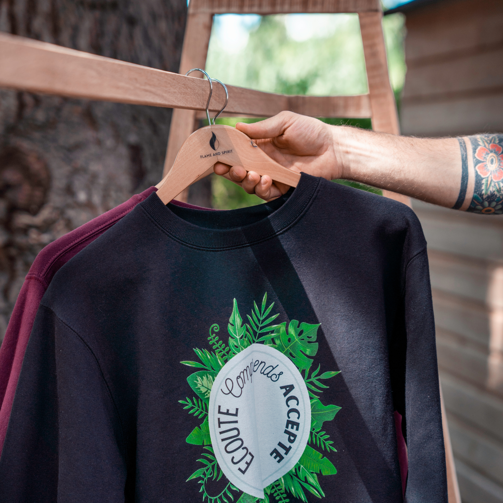 Sweat noir Flame & Spirit suspendu sur un cintre, avec motif feuillage et message "Écoute, comprends, accepte".