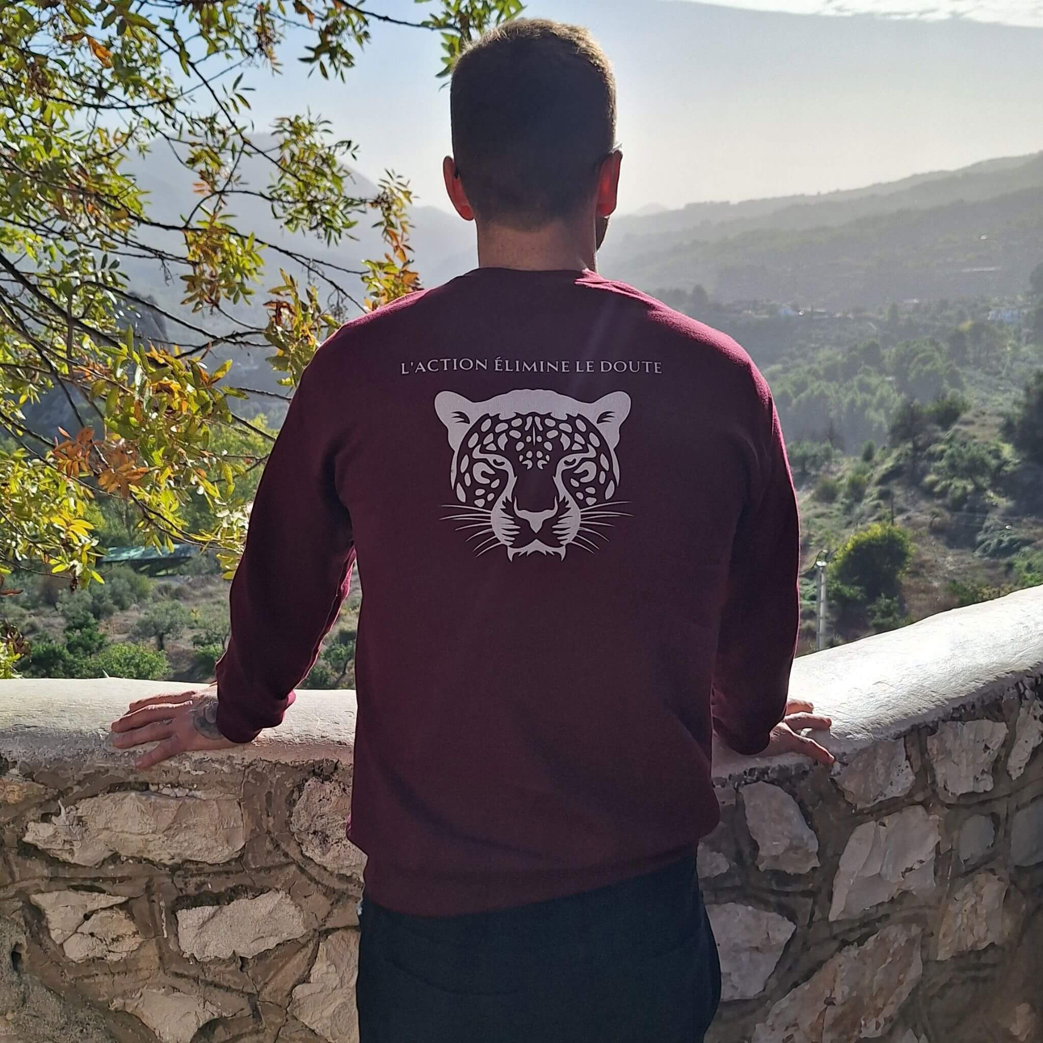 Homme portant un sweat bordeaux Flame & Spirit avec motif tête de félin et message "L’action élimine le doute", face à un paysage naturel.