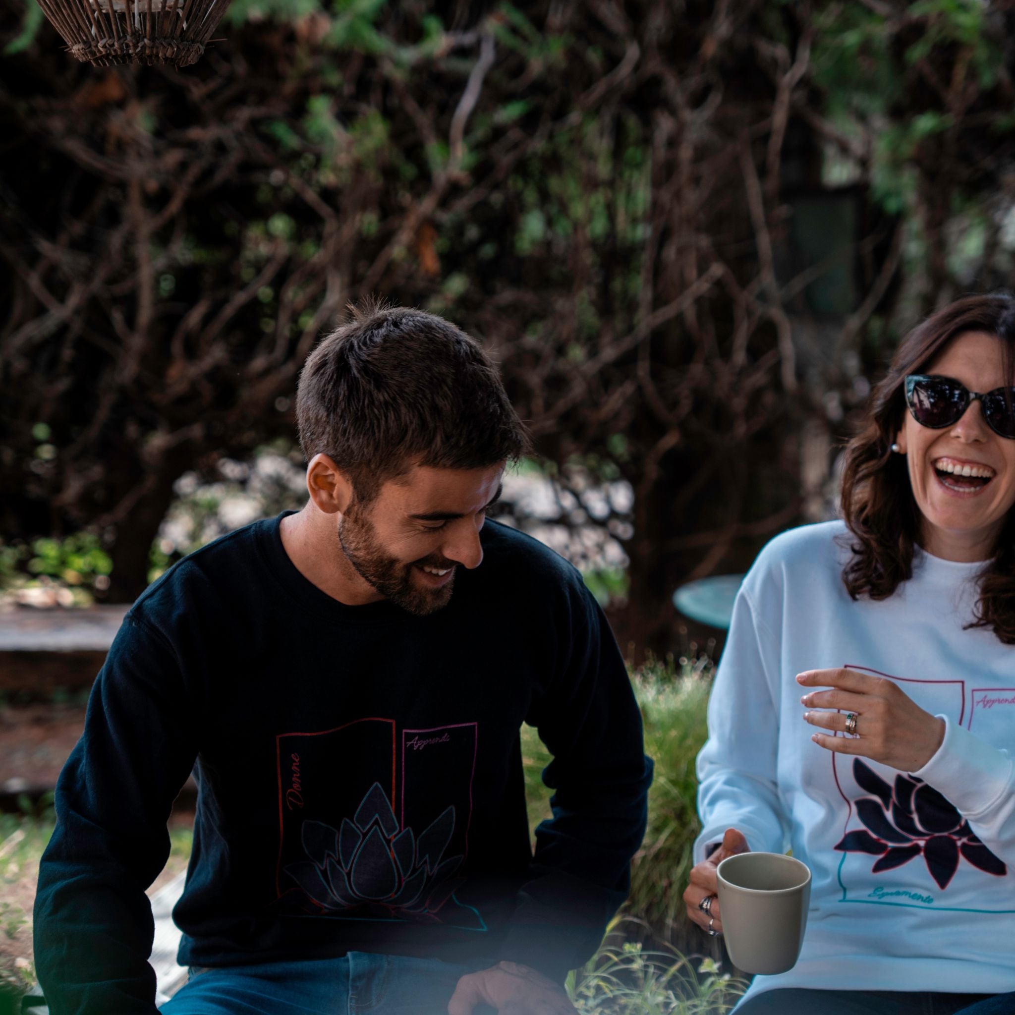 Deux personnes portant des sweats Flame & Spirit avec motif lotus, l’un noir et l’autre blanc, assis en extérieur et souriants.