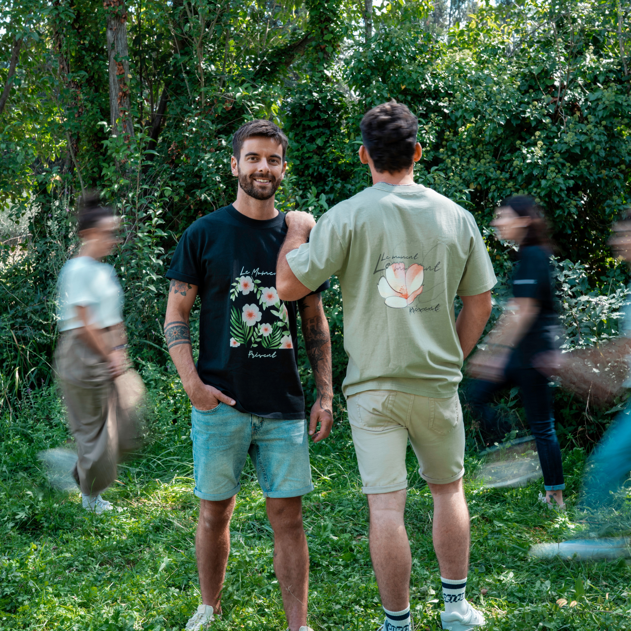 Deux personnes portent des t-shirts Flame & Spirit avec imprimé floral et message "Le moment présent", en extérieur dans un décor naturel.