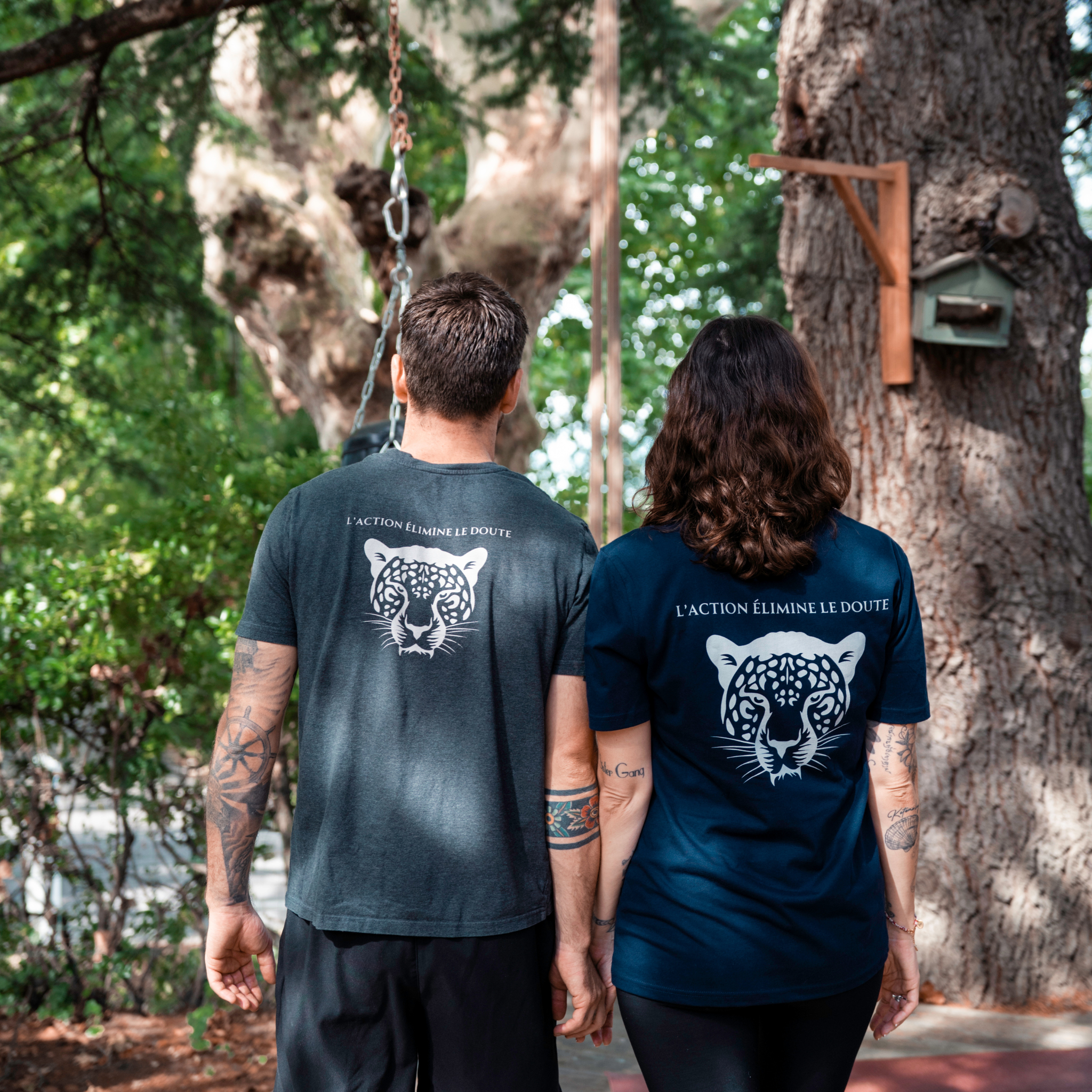 Couple portant les t-shirts Flame & Spirit avec motif tigre et message “L’action illumine la route”, modèles bleu et gris, photo en extérieur.